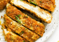Close-up of crispy, baked chicken cutlets sliced into strips on a white plate, garnished with chopped parsley.