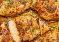 Close-up of juicy, golden-brown Garlic Butter Chicken breasts sprinkled with herbs and spices, one piece sliced to reveal the moist interior, all surrounded by a rich, savory sauce.