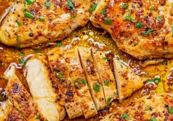Close-up of juicy, golden-brown Garlic Butter Chicken breasts sprinkled with herbs and spices, one piece sliced to reveal the moist interior, all surrounded by a rich, savory sauce.