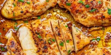 Close-up of juicy, golden-brown Garlic Butter Chicken breasts sprinkled with herbs and spices, one piece sliced to reveal the moist interior, all surrounded by a rich, savory sauce.