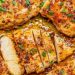 Close-up of juicy, golden-brown Garlic Butter Chicken breasts sprinkled with herbs and spices, one piece sliced to reveal the moist interior, all surrounded by a rich, savory sauce.