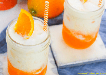 Two mason jars filled with a creamy, orange Dirty Soda, topped with ice, an orange slice, and an orange-striped straw, sit on light coasters with a blue cloth underneath.
