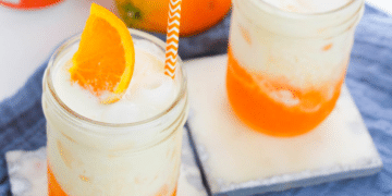 Two mason jars filled with a creamy, orange Dirty Soda, topped with ice, an orange slice, and an orange-striped straw, sit on light coasters with a blue cloth underneath.