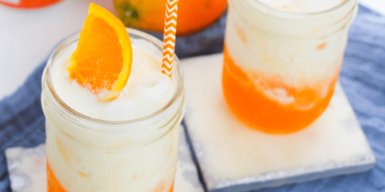 Two mason jars filled with a creamy, orange Dirty Soda, topped with ice, an orange slice, and an orange-striped straw, sit on light coasters with a blue cloth underneath.