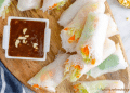 A wooden board with fresh Summer Rolls filled with vegetables and rice noodles, sliced in half, served with a small dish of brown dipping sauce topped with chopped peanuts. A blue and white towel is partially visible on the side.