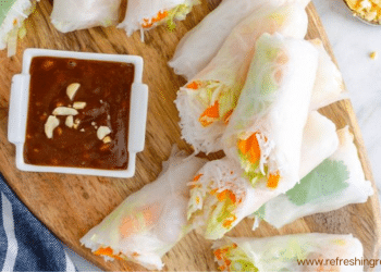 A wooden board with fresh Summer Rolls filled with vegetables and rice noodles, sliced in half, served with a small dish of brown dipping sauce topped with chopped peanuts. A blue and white towel is partially visible on the side.