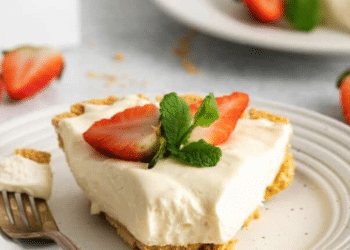 A slice of creamy 3 ingredient no-bake cheesecake garnished with two strawberry halves and a sprig of mint sits on a white plate with a fork. More strawberries and another plate are visible in the background.