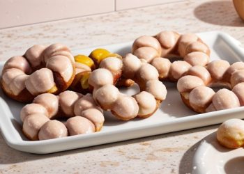 A white rectangular tray holds several glazed, ring-shaped mochi donuts with bubble-like segments, set on a light countertop. A small dish with part of a mochi donut is visible in the corner.
