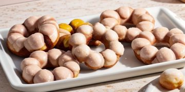 A white rectangular tray holds several glazed, ring-shaped mochi donuts with bubble-like segments, set on a light countertop. A small dish with part of a mochi donut is visible in the corner.