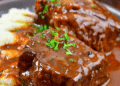 Boneless Beef Short Ribs braised and covered in rich brown gravy, garnished with chopped parsley, served alongside a portion of creamy mashed potatoes on a dark plate.