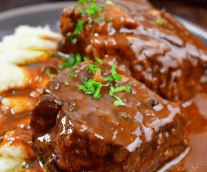 Boneless Beef Short Ribs braised and covered in rich brown gravy, garnished with chopped parsley, served alongside a portion of creamy mashed potatoes on a dark plate.