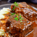 Boneless Beef Short Ribs braised and covered in rich brown gravy, garnished with chopped parsley, served alongside a portion of creamy mashed potatoes on a dark plate.