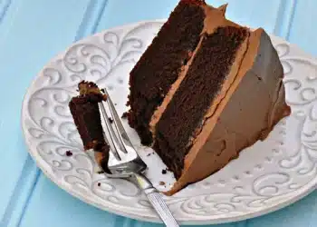 A slice of Beatty's Chocolate Cake with rich chocolate frosting sits on a decorative white plate. A fork rests on the plate with a small bite of cake. The background is a light blue wooden surface.