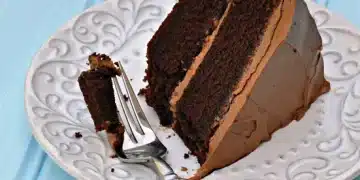 A slice of Beatty's Chocolate Cake with rich chocolate frosting sits on a decorative white plate. A fork rests on the plate with a small bite of cake. The background is a light blue wooden surface.