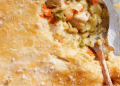 A close-up of a golden, flaky pot pie crust with a spoon scooping out creamy filling made of chicken, peas, carrots, and other vegetables—truly the best comfort food for any occasion.