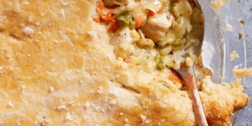 A close-up of a golden, flaky pot pie crust with a spoon scooping out creamy filling made of chicken, peas, carrots, and other vegetables—truly the best comfort food for any occasion.