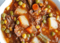 A bowl of old fashioned vegetable beef soup filled with hearty chunks of beef, potatoes, peas, carrots, corn, and green beans in a rich tomato-based broth.