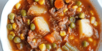 A bowl of old fashioned vegetable beef soup filled with hearty chunks of beef, potatoes, peas, carrots, corn, and green beans in a rich tomato-based broth.