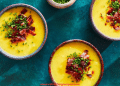 Three bowls of creamy yellow Fall Soup topped with chopped bacon and fresh chives, arranged on a teal surface. A small bowl of chopped chives sits nearby.