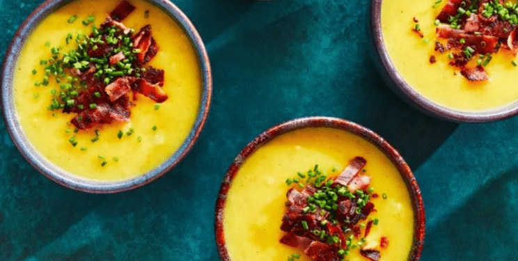 Three bowls of creamy yellow Fall Soup topped with chopped bacon and fresh chives, arranged on a teal surface. A small bowl of chopped chives sits nearby.