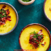 Three bowls of creamy yellow Fall Soup topped with chopped bacon and fresh chives, arranged on a teal surface. A small bowl of chopped chives sits nearby.