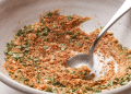 A close-up of a white bowl containing a flavorful meatloaf seasoning recipe—a blend of dry herbs and spices—with a silver spoon partially submerged, ready to elevate your next dish.