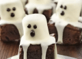 Chocolate mini cakes decorated as ghosts with draped white icing and chocolate dots for eyes and mouths, arranged on a wooden surface—perfect for fun Recipes with 3 Ingredients for your next spooky celebration.