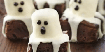 Chocolate mini cakes decorated as ghosts with draped white icing and chocolate dots for eyes and mouths, arranged on a wooden surface—perfect for fun Recipes with 3 Ingredients for your next spooky celebration.