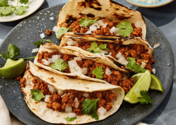 Three tacos filled with seasoned turkey meat, chopped onions, and cilantro are arranged on a gray plate with charred tortillas. This Turkey Tacos Recipe is garnished with lime wedges and fresh cilantro for a burst of flavor.