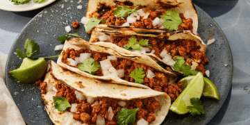 Three tacos filled with seasoned turkey meat, chopped onions, and cilantro are arranged on a gray plate with charred tortillas. This Turkey Tacos Recipe is garnished with lime wedges and fresh cilantro for a burst of flavor.