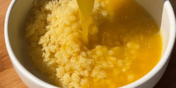 A bowl of cooked rice is topped with a stream of raw beaten egg yolk on a wooden surface, reminiscent of traditional Japanese tamago kake gohan—a comforting dish akin to the warmth found in Italian Penicillin Soup.