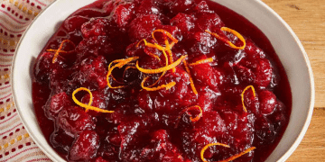 A white bowl filled with chunky cranberry sauce, made from a classic Cranberry Sauce Recipe and garnished with thin strips of orange zest, sits on a wooden surface with a patterned cloth nearby.