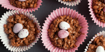 Rice Krispie Buns in pink paper cases, each topped with two speckled candy eggs, are arranged on a dark surface. The website refreshingrecipes.com is visible at the bottom.