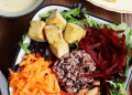 A bowl inspired by the glory bowl recipe, featuring mixed greens, cubed tofu, shredded carrots and beets, wild rice blend, and almonds arranged in sections. A plate and sauce are visible at the edges.