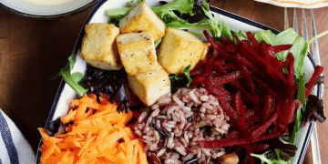 A bowl inspired by the glory bowl recipe, featuring mixed greens, cubed tofu, shredded carrots and beets, wild rice blend, and almonds arranged in sections. A plate and sauce are visible at the edges.