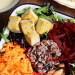 A bowl inspired by the glory bowl recipe, featuring mixed greens, cubed tofu, shredded carrots and beets, wild rice blend, and almonds arranged in sections. A plate and sauce are visible at the edges.