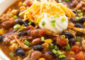 A close-up of a bowl of chicken taco soup with black beans, corn, tomatoes, and shredded chicken—this Taco Soup recipe is topped with sour cream, cheddar cheese, and chopped green onions.