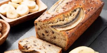 A loaf of angular banana bread with chocolate chips is sliced on a dark surface. A bowl of banana slices and a whole banana are beside the bread.