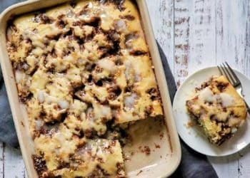 A baked cinnamon crumb cake with icing, perfect for fans of Cinnamon Recipes, sits in a rectangular dish. One square piece is served on a plate with a fork, all arranged on a rustic white wooden surface.