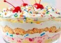 A delightful trifle in a glass bowl with whipped cream, colorful sprinkles, cherries on top, and layers of vanilla pudding, cake, pink cream, and funfetti for a festive touch.