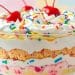 A delightful trifle in a glass bowl with whipped cream, colorful sprinkles, cherries on top, and layers of vanilla pudding, cake, pink cream, and funfetti for a festive touch.