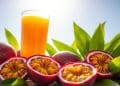 A glass of orange juice is surrounded by whole and halved passion fruits with yellow pulp, resting on green leaves under bright sunlight against a clear blue sky, capturing the essence of refreshing Passion Fruit Juice.