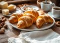 A plate of golden croissants made from a classic gipfeli recipe sits on a wooden table, surrounded by whole almonds, butter, two cups of coffee, and a folded white napkin in a cozy, sunlit setting.