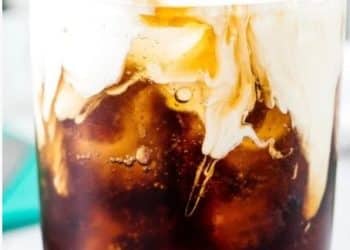 A close-up of a glass filled with iced coffee, topped with a layer of cream swirling into the dark liquid, creating a marbled effect reminiscent of a Dirty Soda fusion.