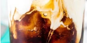 A close-up of a glass filled with iced coffee, topped with a layer of cream swirling into the dark liquid, creating a marbled effect reminiscent of a Dirty Soda fusion.