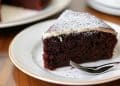 A slice of rich chocolate cake, inspired by Beatty's chocolate cake recipe, with a dusting of powdered sugar and a layer of white icing on top, served on a white plate with a fork.