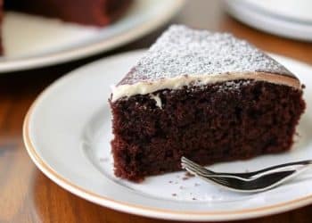 A slice of rich chocolate cake, inspired by Beatty's chocolate cake recipe, with a dusting of powdered sugar and a layer of white icing on top, served on a white plate with a fork.