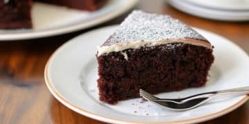 A slice of rich chocolate cake, inspired by Beatty's chocolate cake recipe, with a dusting of powdered sugar and a layer of white icing on top, served on a white plate with a fork.