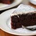 A slice of rich chocolate cake, inspired by Beatty's chocolate cake recipe, with a dusting of powdered sugar and a layer of white icing on top, served on a white plate with a fork.