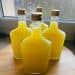 Four square glass bottles filled with bright yellow liquid, crafted from a traditional limoncello recipe and sealed with gold screw caps, are arranged on a wooden surface near a window.